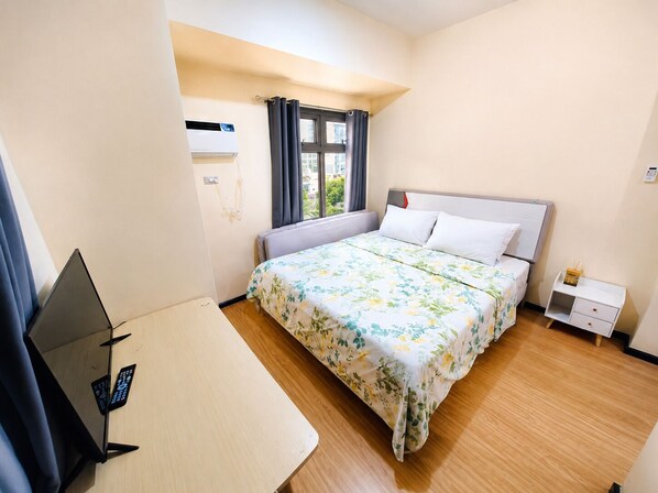 A hotel room with a bed, a desk, a chair, a window with curtains, and an air conditioning unit.
