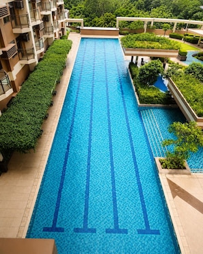 A long swimming pool with clear blue water, surrounded by greenery and a building with balconies.