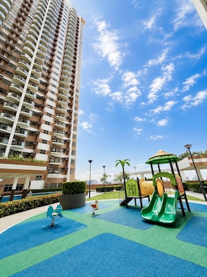 Ein Spielplatz mit Rutsche und Schaukeln, umgeben von einem Hochhaus und blauem Himmel.