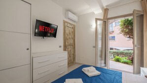 Ein Schlafzimmer mit blauer Bettdecke, einem weißen Schminktisch und einem Fernseher, der 'NETFLIX' anzeigt, sowie Blick auf den Außenbalkon.