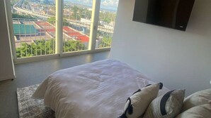 Ein Schlafzimmer mit einem großen Bett, einem flachbildfernseher an der Wand und Blick auf eine Stadt durch große Fenster.