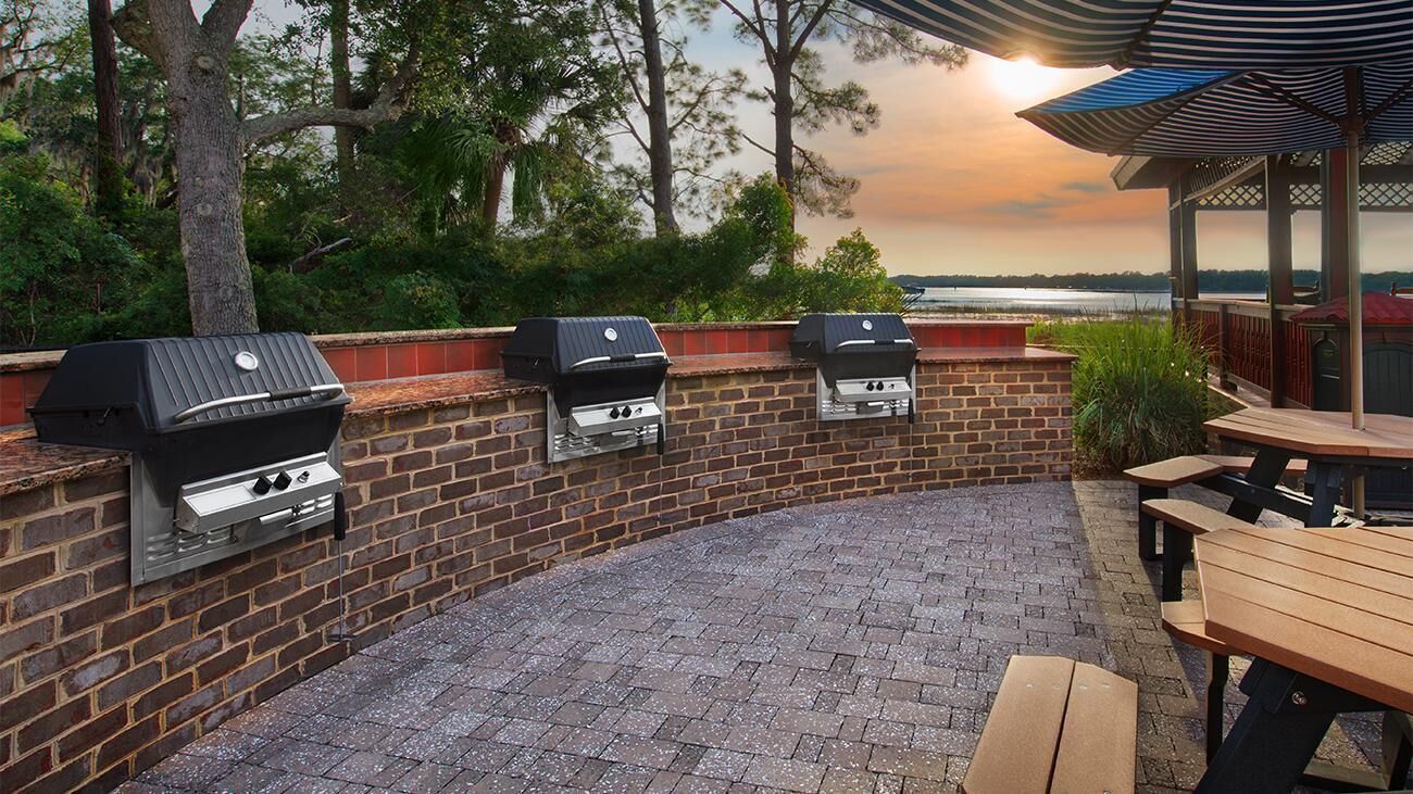 A brick patio with three barbecue grills, a wooden table, and a covered seating area.