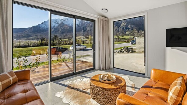 A modern living room with large sliding glass doors, leather sofas, a wooden coffee table, and a view of mountains and parked cars.