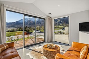 A modern living room with large sliding glass doors, leather sofas, a wooden coffee table, and a view of mountains and parked cars. - Remarkables Retreat, close to Ski Fields! (Hanleys Farm)