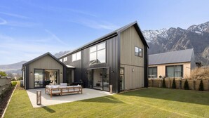 A modern two-story house with a large patio area, a wooden deck, and a view of mountains in the background.