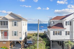 A coastal residential area with two houses, one with a balcony and a red door, and the other with a red roof. A boat is visible on the water in the background. - Grand Coastal Getaways – 7BR Pool & Beach Path, Perfect for Making Memories! (Murrells Inlet)