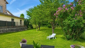 Un patio con una mesa, sillas y una tumbona. Hay plantas en macetas y un pequeño jardín.