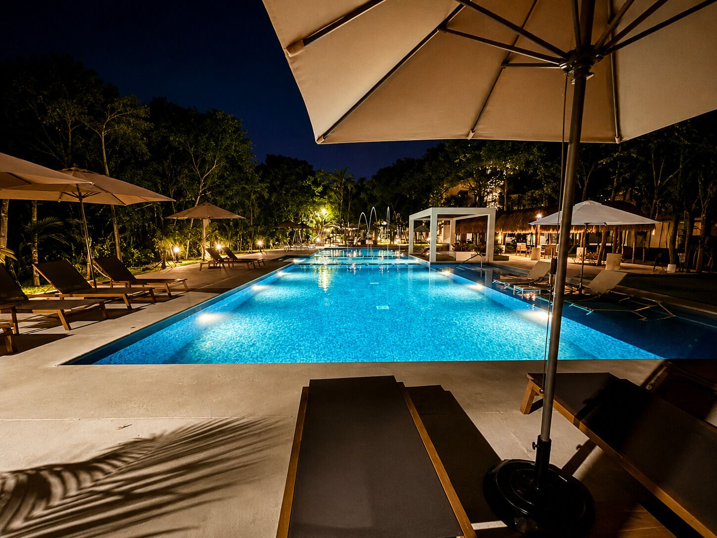 A swimming pool with lounge chairs and umbrellas, surrounded by trees and night lighting.