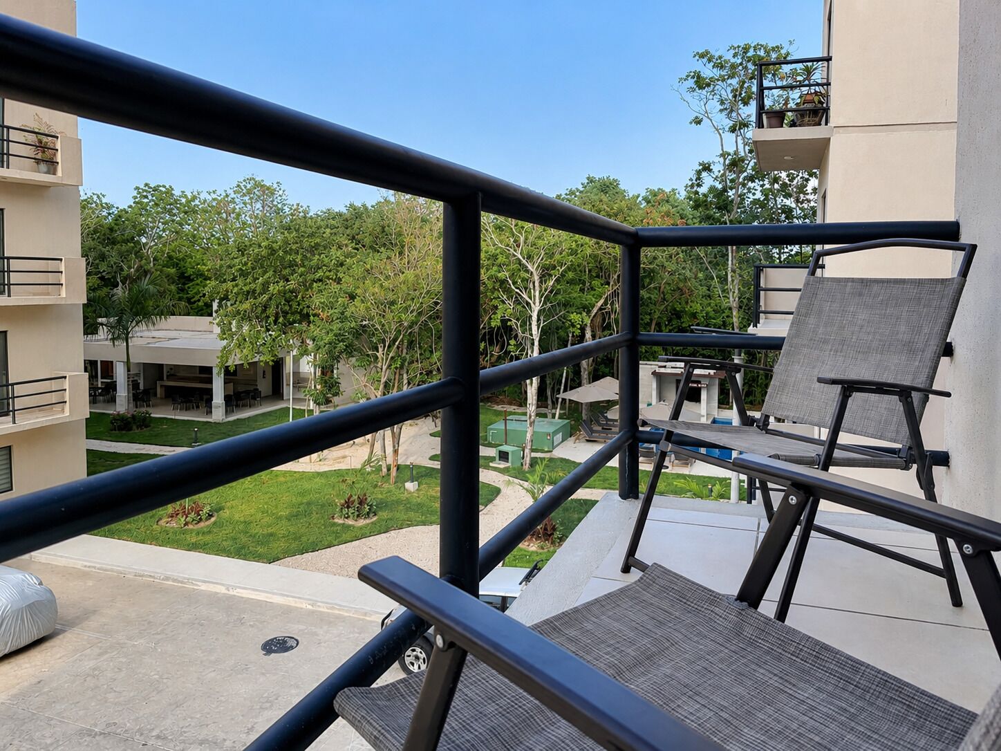 A balcony with outdoor furniture, a view of a courtyard with greenery and buildings, and a clear sky.