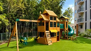 Een houten speelhuisje met een glijbaan en klimmuur, omringd door een omheinde tuin met kunstgras.