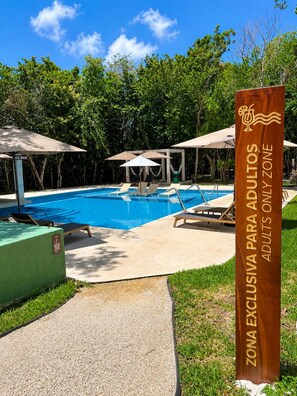 A clear blue pool with lounge chairs and umbrellas, surrounded by greenery. A sign indicates an adults-only zone.