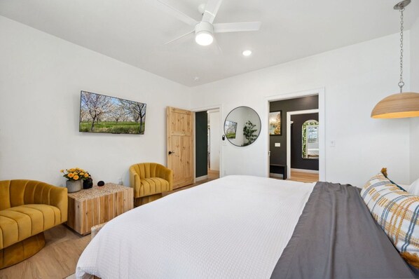 A modern bedroom with a large bed, a ceiling fan, a round mirror, and a painting on the wall.