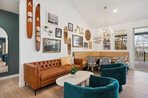 A living room with a brown sofa, teal armchairs, a coffee table, and a dining area with chairs and a table.