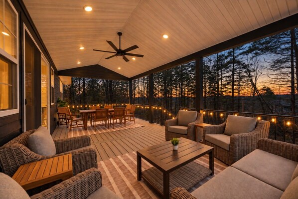 A covered outdoor patio with wicker furniture, a ceiling fan, string lights, and a view of trees at dusk.