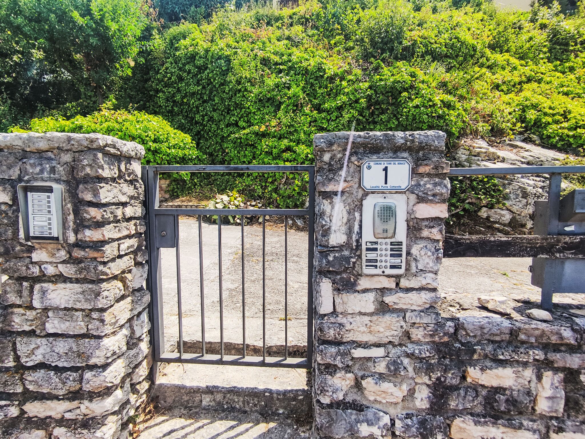 Une porte en pierre surmontée d’un portail en métal, une plaque indiquant « COMUNE DI TORINO BENACO 1 », et une cabine téléphonique.