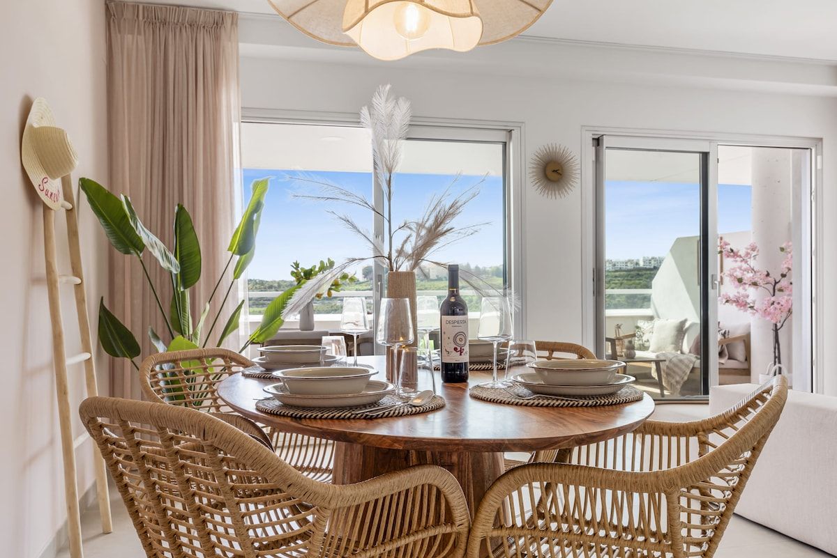 A dining area with a round wooden table, wicker chairs, and a bottle of wine.