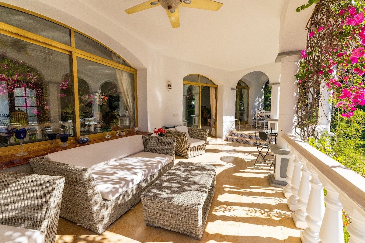 A sunlit outdoor seating area with wicker furniture, a ceiling fan, and a view of the garden.
