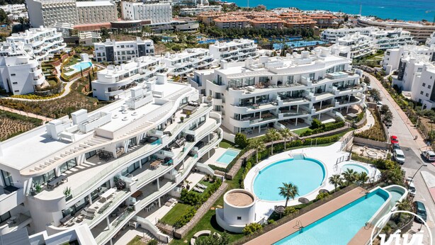A modern resort with multiple buildings, a central swimming pool, and a view of the sea.