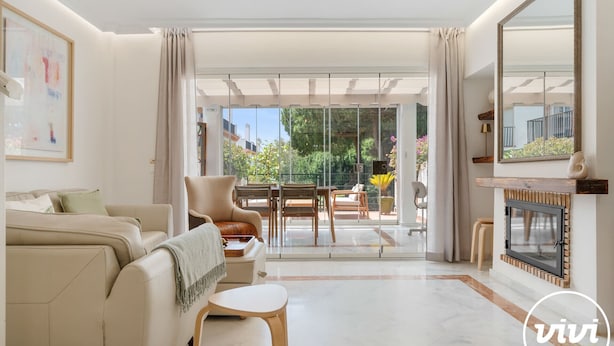 A modern living room with a sofa, a small table, and a fireplace. There is a large window with curtains, and a view of greenery outside. - Lochloosa | Pool - terrace - golf (Marbella)