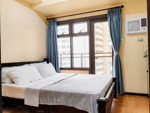 A hotel room with a bed, a window with curtains, a wall-mounted air conditioner, and a view of a building outside.