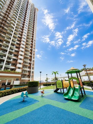 Ein Spielplatz mit Rutsche und Schaukeln, umgeben von einem Hochhaus und blauem Himmel.