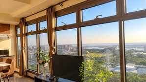A modern hotel room with a large window, a flat-screen TV, a dining area, and a plant.