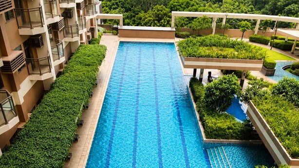 A long swimming pool with clear blue water, surrounded by greenery and a building with balconies.