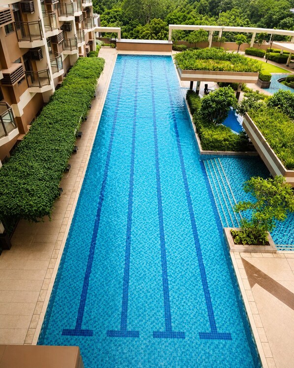 Una piscina larga con agua azul cristalina, rodeada de follaje y un edificio con balcones.