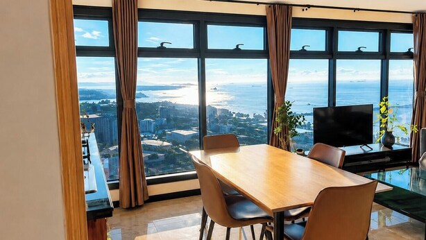 Un comedor con una mesa grande y sillas, televisión y vistas a un paisaje urbano y un cuerpo de agua a través de amplios ventanales.