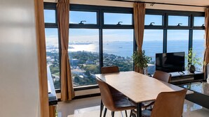 Un comedor moderno con una mesa grande y sillas, un televisor de pantalla plana, y vistas a un cuerpo de agua y al paisaje urbano a través de amplias ventanas.