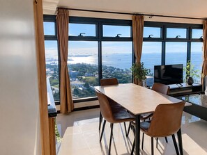 A modern dining area with a large table and chairs, a flat-screen TV, and a view of a body of water and cityscape through large windows.