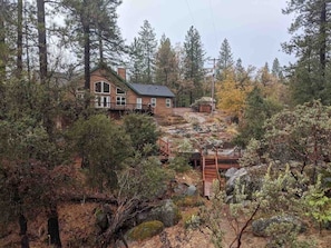 A cabin surrounded by dense forest, with a wooden deck and a small staircase leading to the entrance.