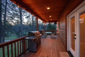 A covered deck with a grill, outdoor table, and chairs, overlooking a forested area.