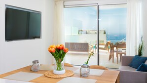 A modern living room with a flat-screen TV, a wooden dining table with placemats, a vase of tulips, and a view of a balcony with outdoor furniture.