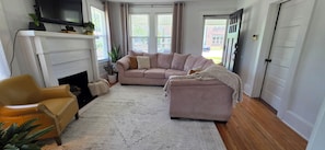Sala de estar com lareira, sofá seccional rosa, poltrona amarela e uma janela grande com persianas.