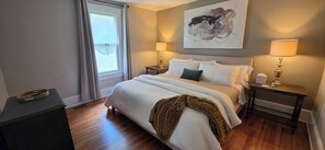 Quarto com cama, duas mesas de cabeceira, janela com cortinas, uma obra de arte na parede e abajur no lado direito.