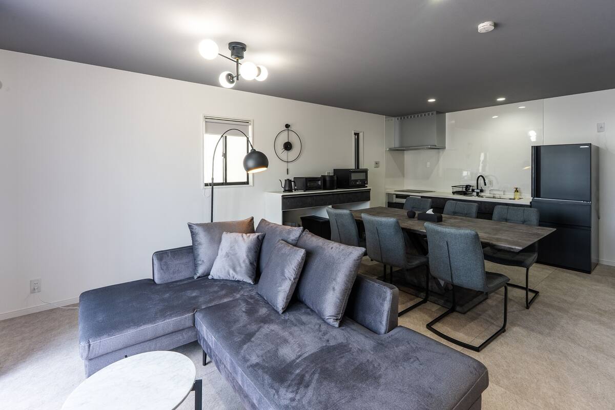 A modern living room with a grey sectional sofa, a dining area with grey chairs, a kitchen with white cabinets, and a large flat-screen TV.