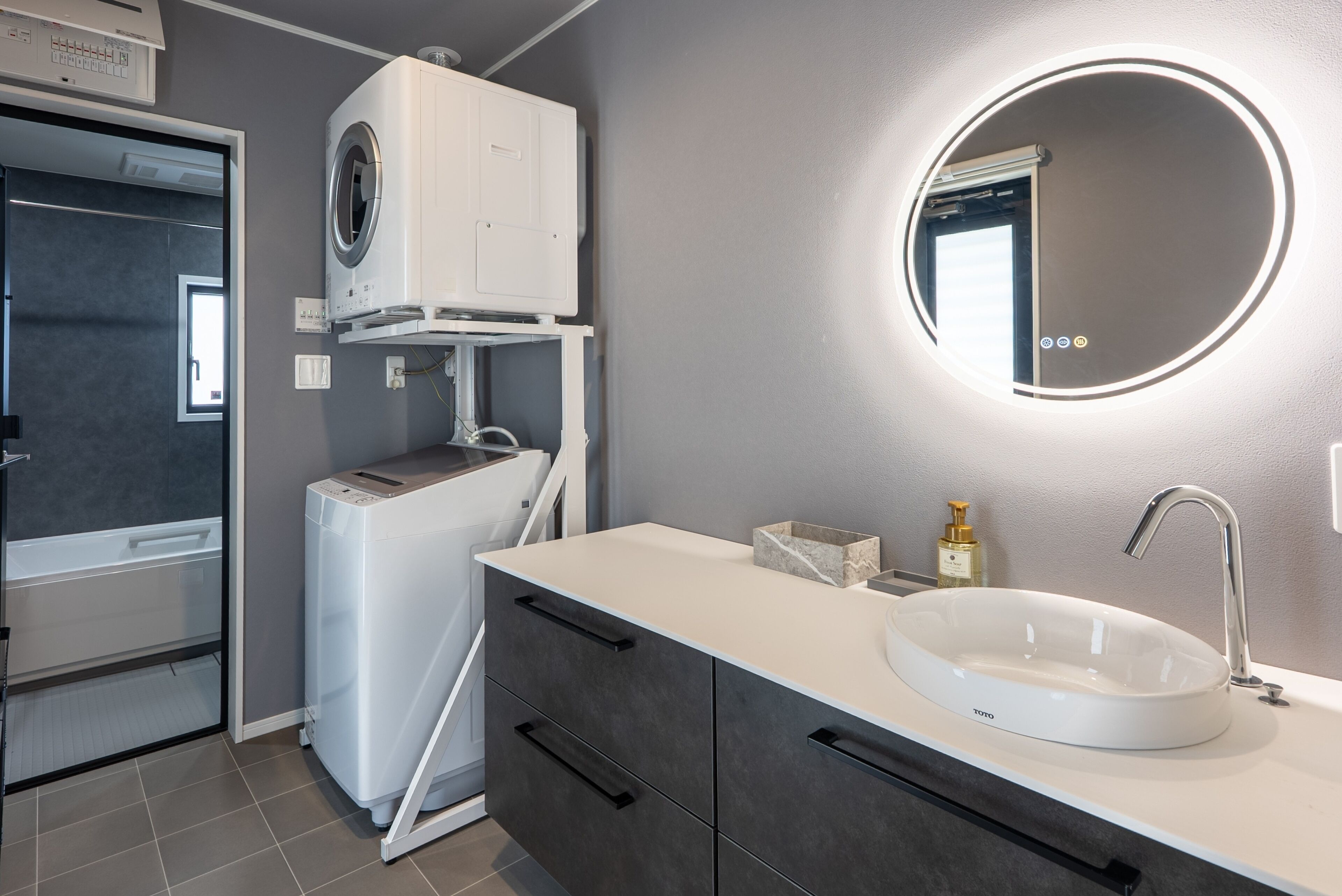 Une salle de bain moderne équipée d’un lave-linge intégré, d’un miroir rond avec éclairage, d’un lavabo et d’un meuble de rangement.