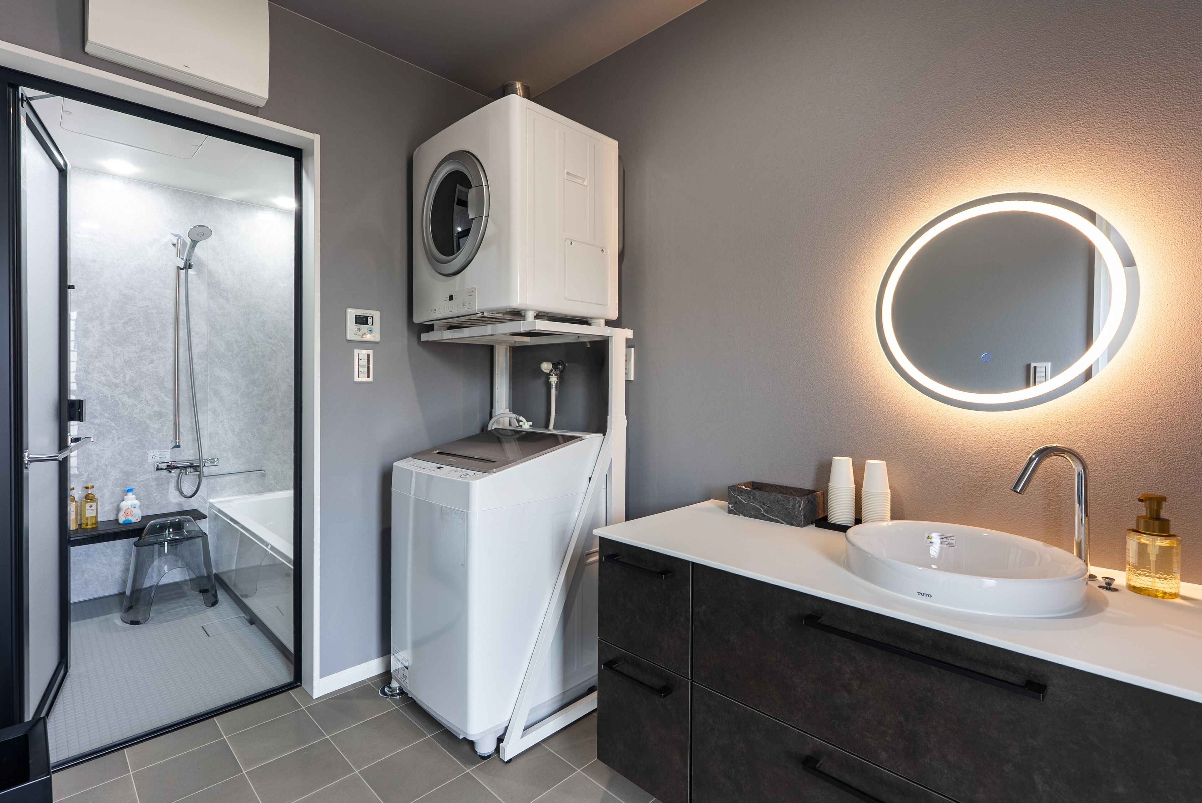 A modern bathroom with a built-in washing machine, a round mirror with a light, and a sink with a faucet.