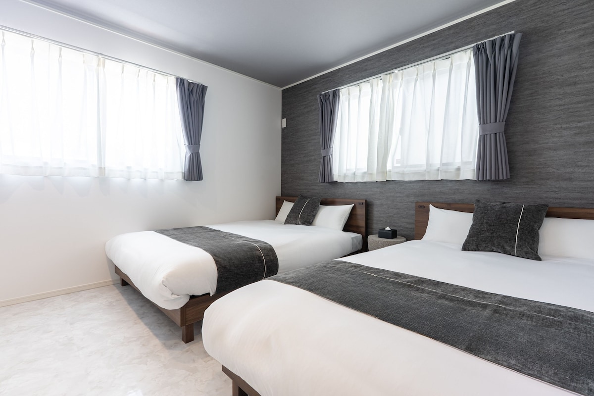 Habitación de hotel con dos camas, ventanas amplias con cortinas, pared gris y cabecero de madera.