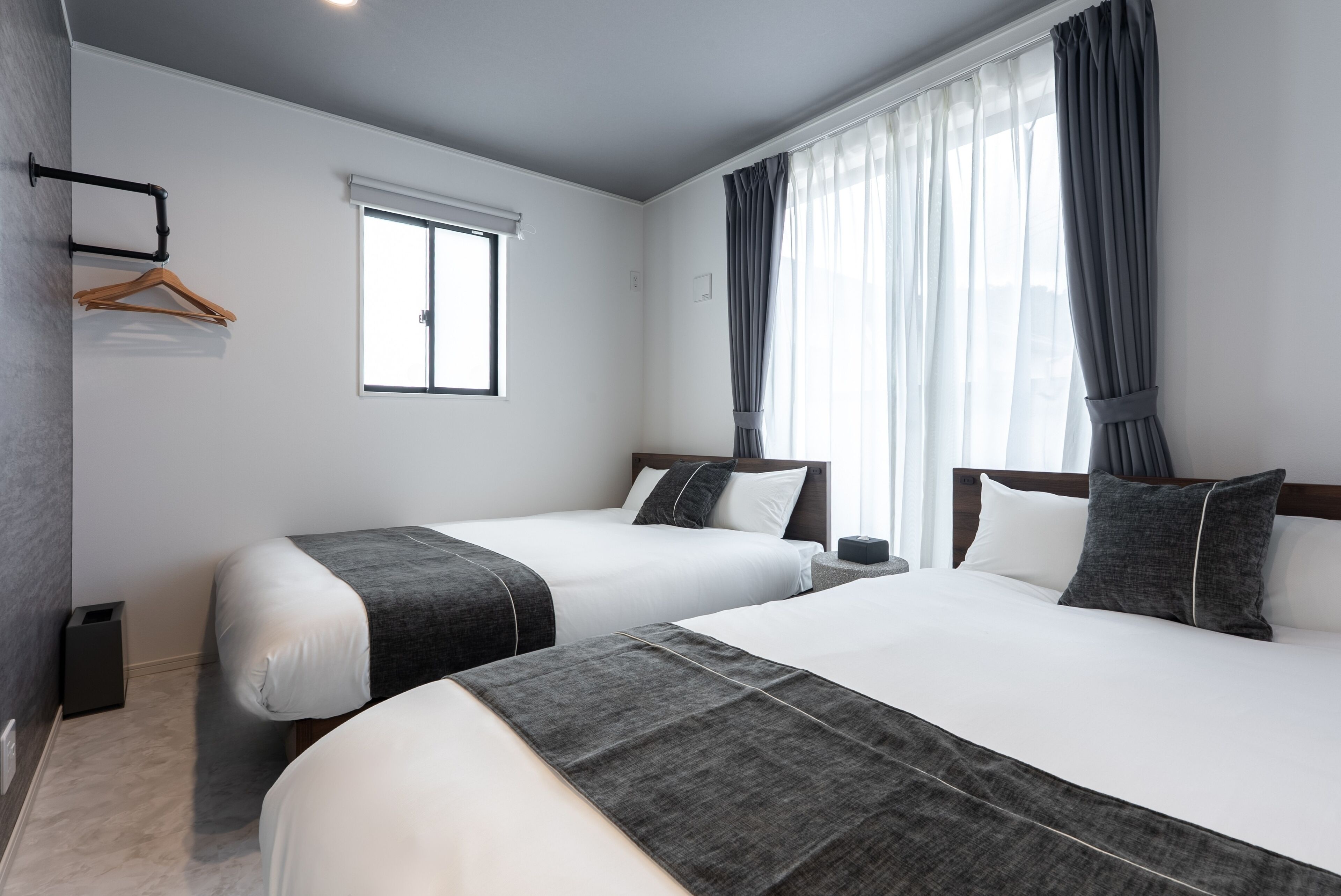 A hotel room with two beds, a window with blinds, a hanging coat rack, and a bedside table with a lamp.