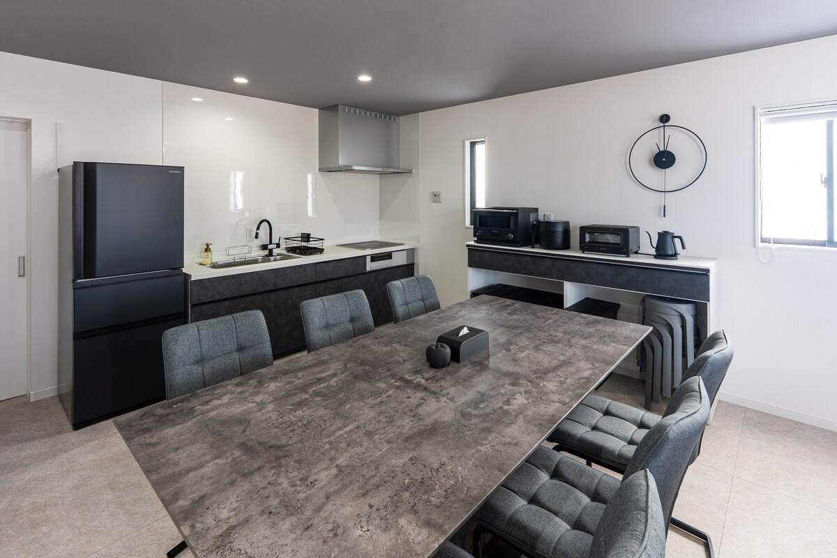 Una cocina moderna con una isla grande, un comedor con una mesa larga y sillas, y una zona de estar compacta con kitchenette.