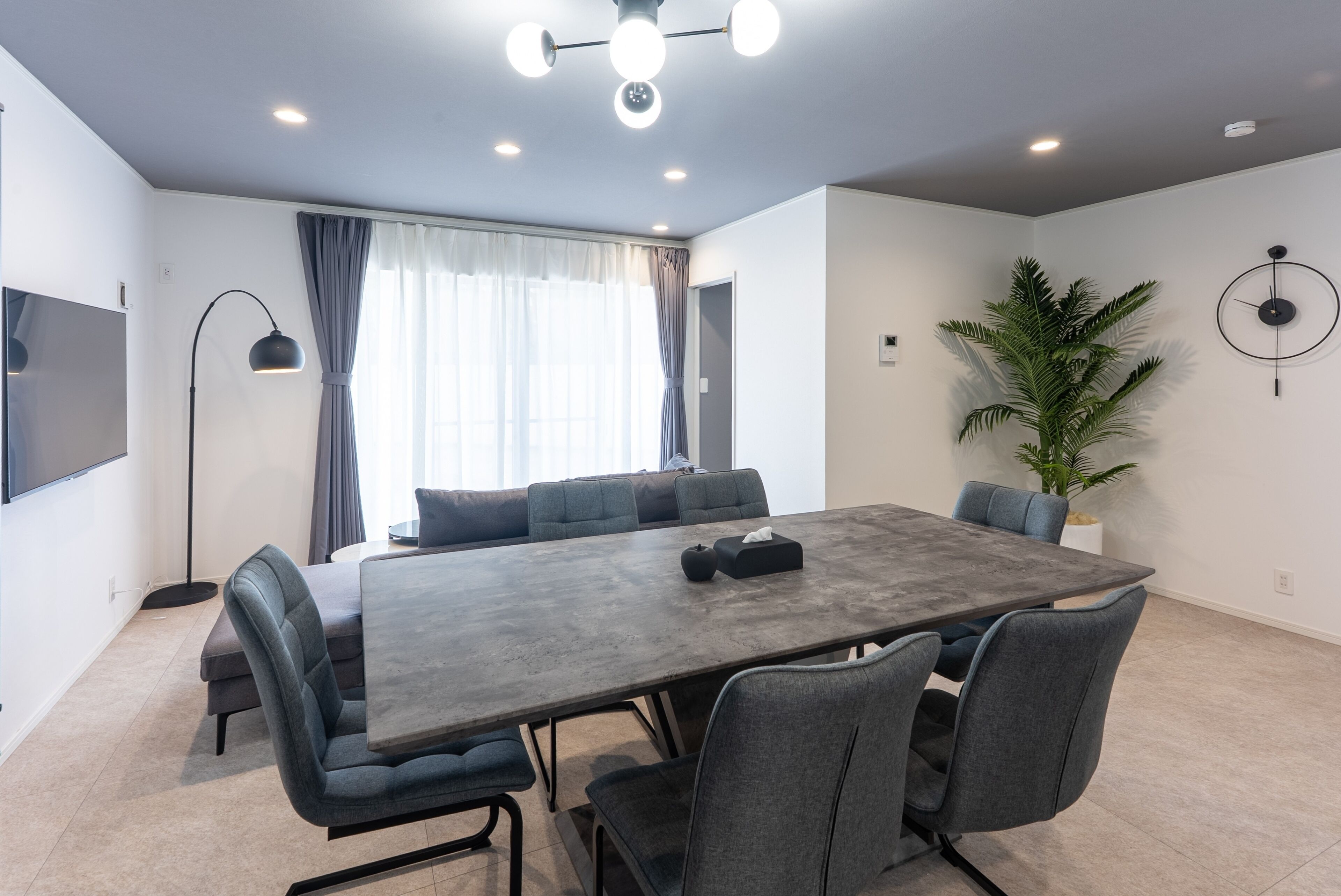A modern meeting room with a large table, grey upholstered chairs, a floor lamp, a potted plant, and a wall-mounted clock.