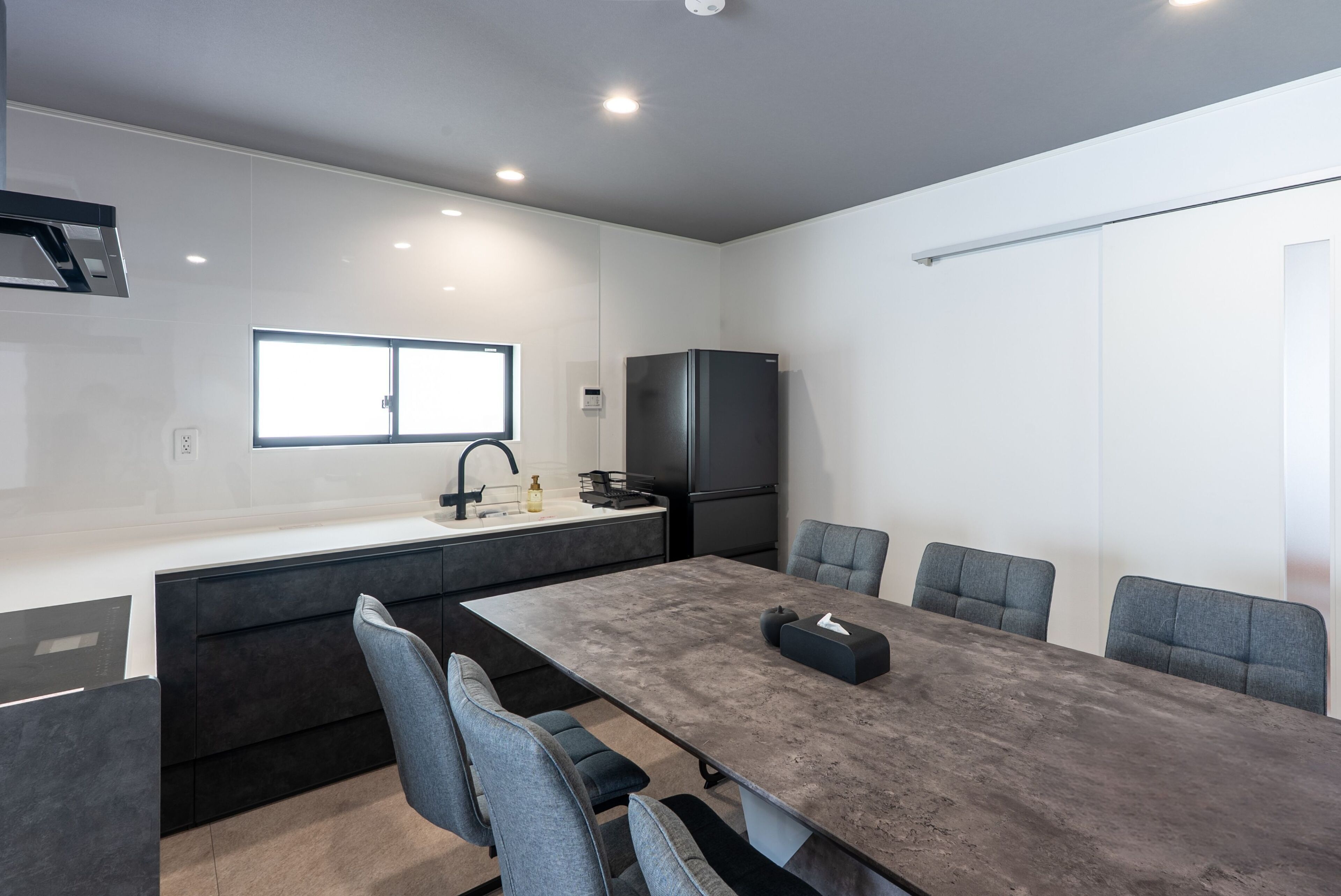Une cuisine moderne avec un coin repas, comprenant une grande table, des chaises grises, un réfrigérateur noir et une fenêtre.
