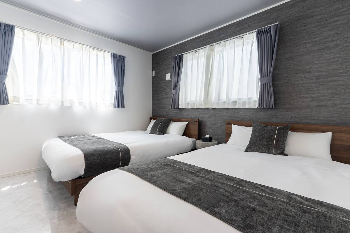 A modern hotel room with two beds, grey and white bedding, and large windows with curtains.