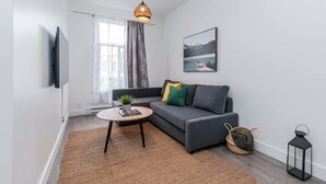 Een moderne woonkamer met een bank, een rond bijzettafeltje, een televisie, een hanglamp, een ingelijste foto en een raam met gordijnen.
