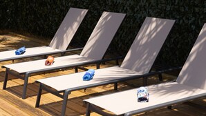 Una hilera de tumbonas con toallas, ubicadas en una terraza de madera con un seto al fondo.