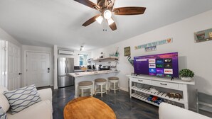 Una cocina moderna con una isla central, un comedor con mesa de madera y una sala de estar con sofá y televisor mostrando varios servicios de streaming.