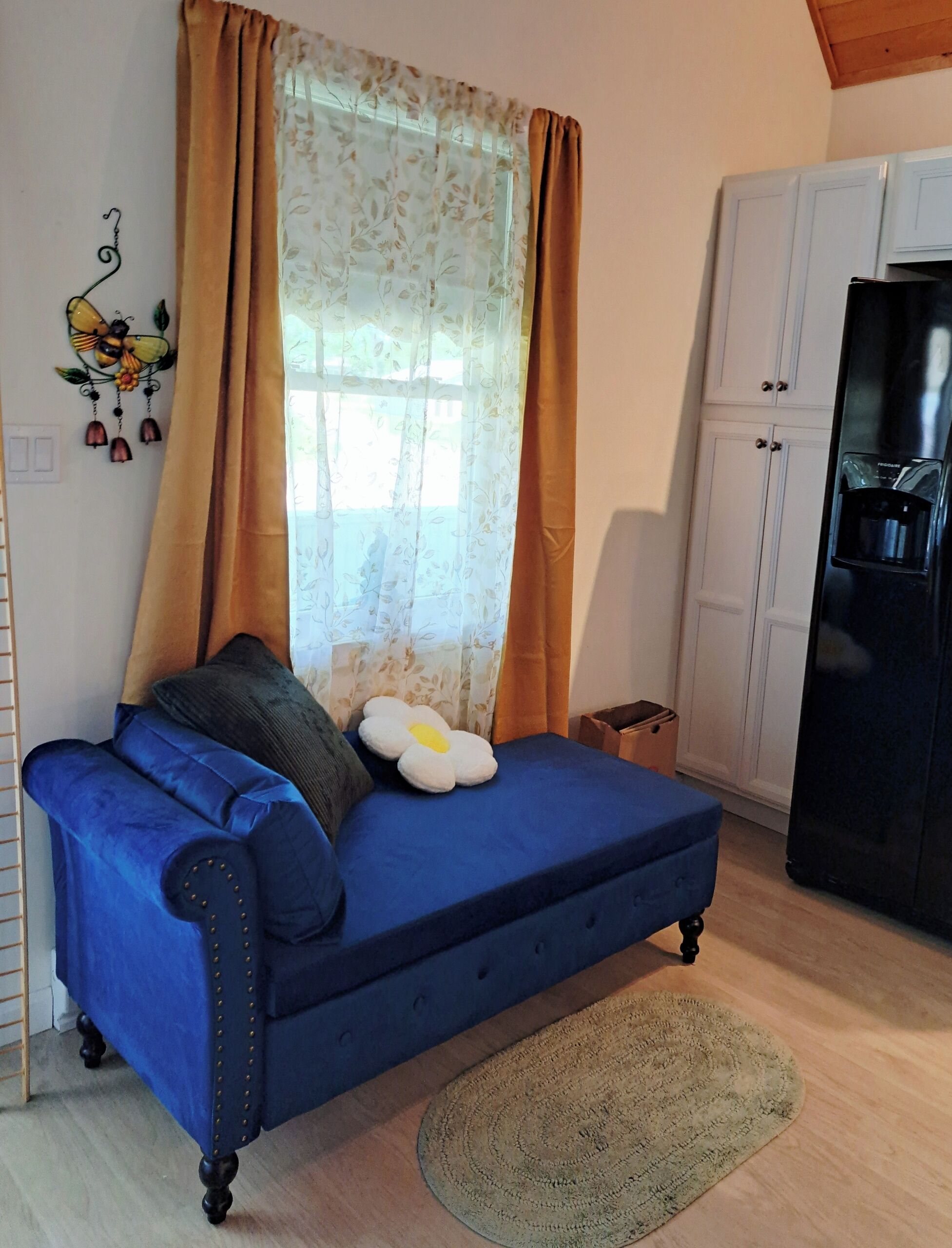 Une pièce avec un canapé bleu, une décoration murale, une fenêtre avec des rideaux, un réfrigérateur et des placards.