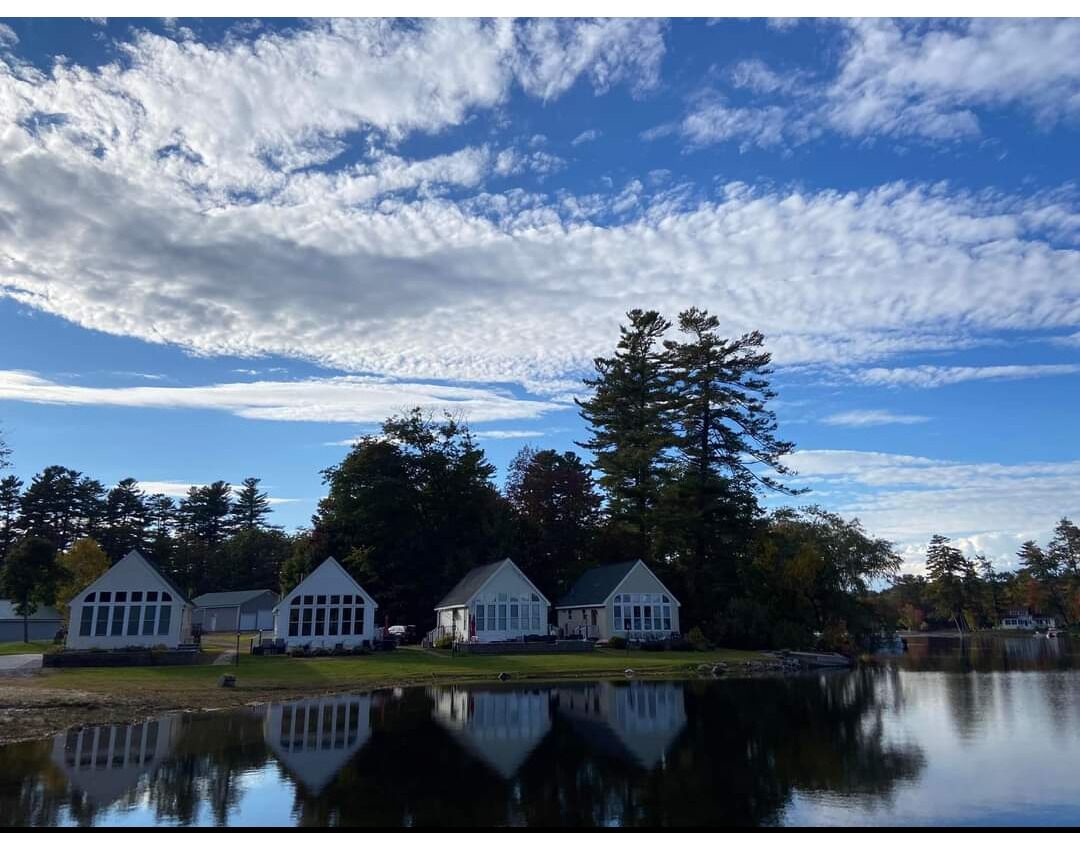 A lakeside property with three white buildings, a large tree, and a calm lake reflecting the structures.